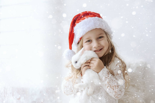 linda menina em um vestido branco com um pequeno coelho branco em um chapéu de santa claus
 - Foto, Imagem