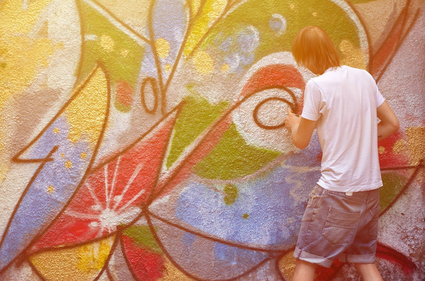 Foto en el proceso de dibujar un patrón de graffiti en una vieja pared de hormigón. Joven rubio de pelo largo dibuja un dibujo abstracto de diferentes colores. Arte callejero y concepto de vandalismo
 - Foto, Imagen