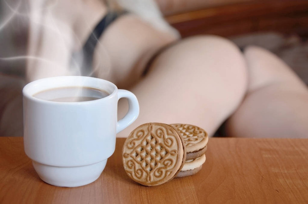 eine Tasse heißen Kaffee und runde Kekse mit der Silhouette eines sexy Mädchens in schwarzer Unterwäsche im Hintergrund auf einem Bett. Schwerpunkt Priorität. kleine weiße Kaffeetasse mit Dampf und braunen Keksen  - Foto, Bild