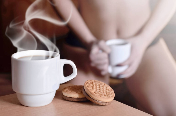 Una taza de café caliente y galletas redondas con una silueta de una chica sexy en ropa interior negra en el fondo en una cama. Enfoque prioridad de primer plano. Pequeña taza de café blanco con vapor y galletas marrones
  - Foto, imagen