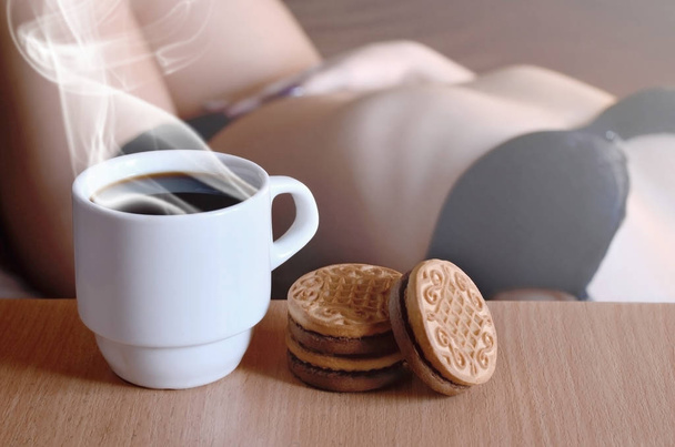 Чашка гарячої кави і кругле печиво з силуетом сексуальної дівчини в чорній білизні на задньому плані на ліжку. Пріоритет переднього плану фокусу. Маленька біла чашка кави з паром і коричневим печивом
  - Фото, зображення