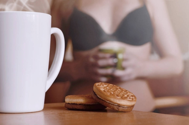 Šálek horké kávy a kulaté sušenky s silueta sexy dívka v černém prádle v pozadí na posteli. Priorita popředí zaměření. Malý bílý šálek s párou a hnědé sušenky  - Fotografie, Obrázek