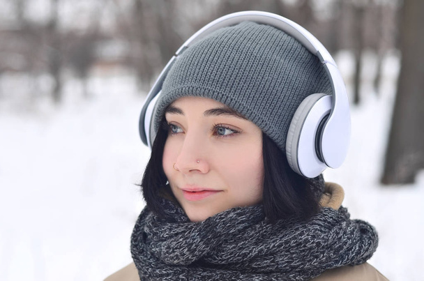 Портрет довольно привлекательной кавказской девушки с пирсингом хипстерского зимнего парка. Она слушает музыку в наушниках. Молодежная мода. Зимнее веселье
. - Фото, изображение