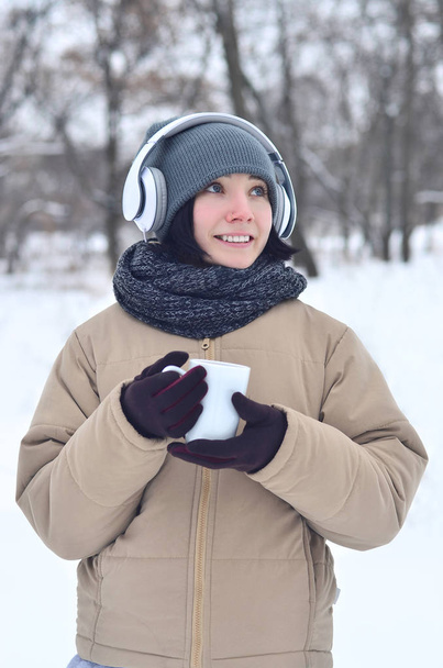 schöne gepiercte brünette kaukasische junge Frau lächelt, während sie Musik auf großen weißen Kopfhörern Musik hört und eine Tasse Kaffee im Winter Park oder Wald trinkt - Foto, Bild