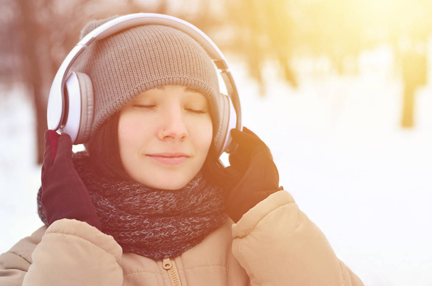 Beauty girl in winter forest. Beautiful young brunette caucasian pierced woman listening to music with big white earphones in fashionable brown coat and warm black scarf - Photo, Image