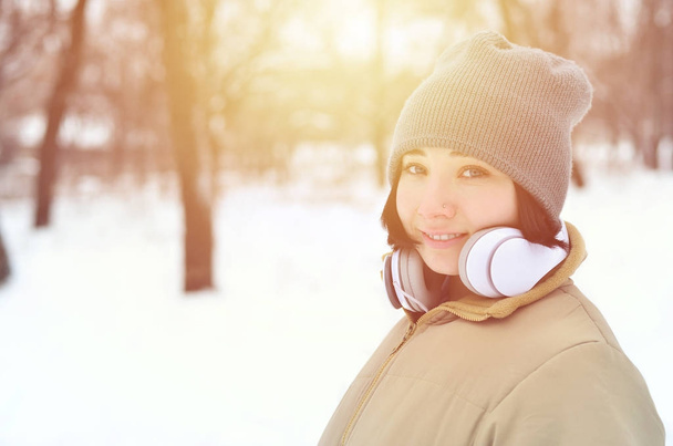Ομορφιά κορίτσι στο δάσος του χειμώνα. Όμορφη νεαρή μελαχρινή Καυκάσιος τρυπημένα γυναίκα να ακούτε μουσική με μεγάλα λευκά ακουστικά σε μοντέρνα καφέ παλτό και ζεστά κασκόλ μαύρο - Φωτογραφία, εικόνα