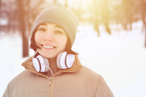 Mooie jonge meisje draag warme winter oorkappen en sjaal. Leuke brunette vrouwelijke model wandeling buiten met koptelefoon in koud weer. Glimlachend gelukkig model poseren. Goede vakantie weer - Foto, afbeelding