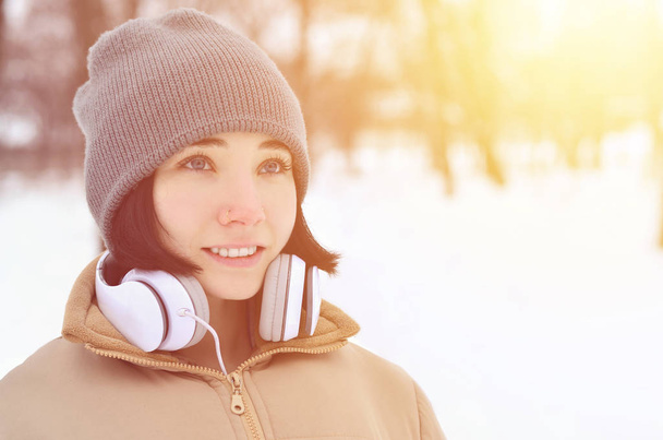 Красавица в зимнем лесу. Красивая юная брюнетка кавказская женщина с пирсингом слушает музыку с большими белыми наушниками в модном коричневом пальто и теплым черным шарфом
 - Фото, изображение