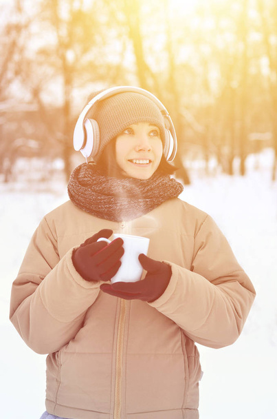 Winter portrait of pretty attractive caucasian pierced girl in snowy wood. She is listening to music on big white headphones with coffee cup in her hands. Walks in the open air - Photo, Image