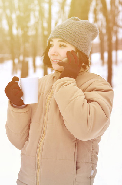 Porträt einer hübschen kaukasischen gepiercten Hipsterin im Winterpark. Sie spricht auf einem modernen Smartphone und trinkt Kaffee. Jugend-Straßenmode. Spaziergänge im Freien - Foto, Bild