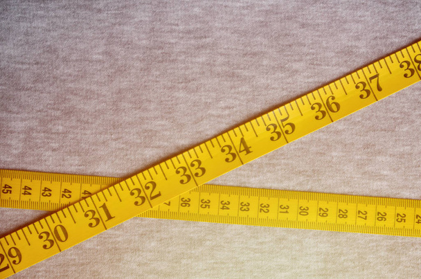 Измерительная лента желтого цвета с числовыми индикаторами в виде сантиметров или дюймов лежит на серой трикотажной ткани. Фон изображения о шитье спортивной одежды для людей, которые похудения
 - Фото, изображение