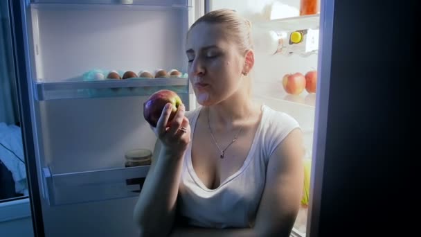 Krásná mladá žena jíst čerstvé jablko na kuchyň v noci - Záběry, video