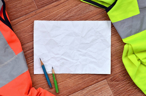 Una hoja de papel arrugada con dos lápices rodeados de uniformes de trabajo verdes y naranjas. Naturaleza muerta asociada a las obras de reparación, ferrocarril o fontanería
 - Foto, Imagen