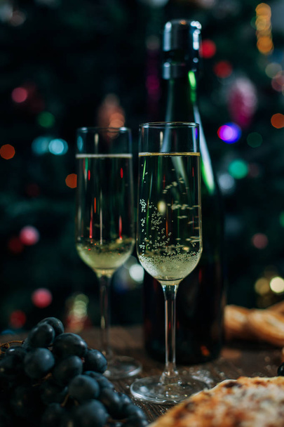 ένα εορταστικό πρωτοχρονιάτικο τραπέζι, κέικ τυριού με μανταρίνια, σταφύλια, σοκολάτα, μπισκότα, σαμπάνια και δυο ποτήρια με φόντο ένα στολισμένο χριστουγεννιάτικο δέντρο με φώτα - Φωτογραφία, εικόνα