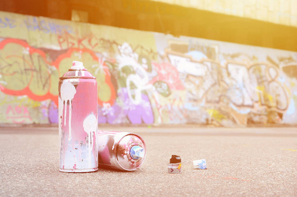 Several used spray cans with pink and white paint and caps for spraying paint under pressure is lies on the asphalt near the painted wall in colored graffiti drawings - 写真・画像