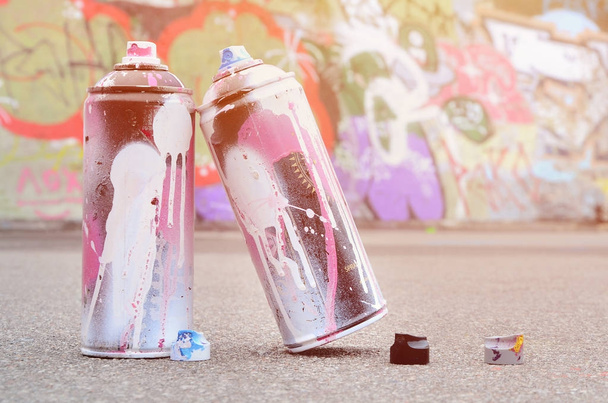 Several used spray cans with pink and white paint and caps for spraying paint under pressure is lies on the asphalt near the painted wall in colored graffiti drawings - Photo, Image