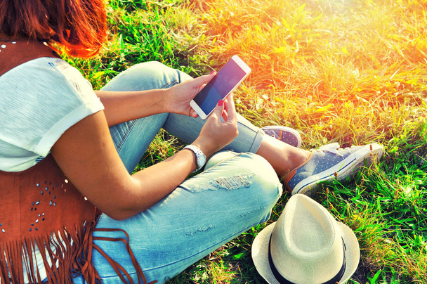 Relaxation with a mobile. Break time. Young woman using cellphone and sitting on the grass wearing jeans, vest of suede leather, white skirt and hat. Sunny  day - Photo, Image