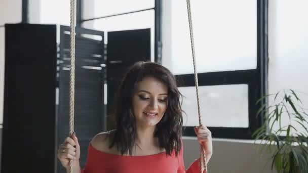 A pregnant woman in a long red dress rides a swing and looks out the window. Smiling pregnant girl on a wooden swing. The concept of pregnancy - soon mother, motherhood, happiness, family - Video