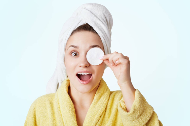 Portrait of excited female with towel on head, covers eye with cotton wheel, demonstrates her care about beauty, prepares for date, takes shower and do make up, isolated over white background - Φωτογραφία, εικόνα