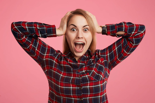 Frightened shocked young female opens mouth widely and exclaims in terror, doesn`t anticipate to see her phobia, keeps hands on head, poses against pink background in checkered stylish shirt - Фото, изображение