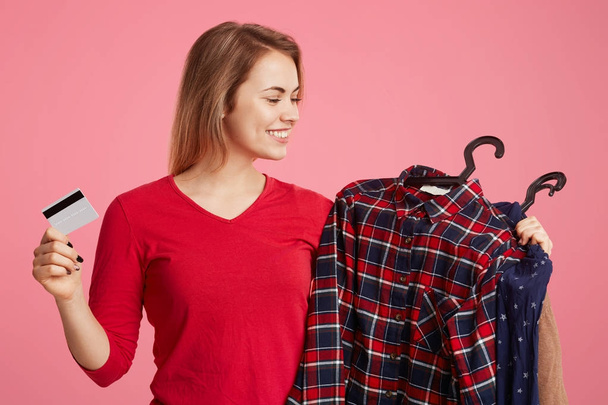 Cheerful smiling female makes successful purchases, looks at her new clothes, holds plastic card as going to pay for outfit, poses against pink background. Woman shopaholic rejoices going shopping - Φωτογραφία, εικόνα