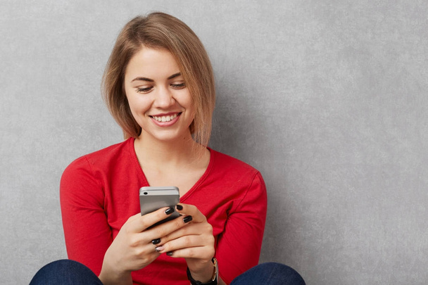 Happy smiling young woman in red sweater holds smart phone and messages online, pleased to recieve text message, sits on floor, isolated over grey background with copy space. Technology concept - Foto, Bild