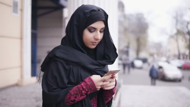 Young attractive woman wearing hijab standing in the street, typing a message on her mobile phone. Slowmotion shot - Кадры, видео