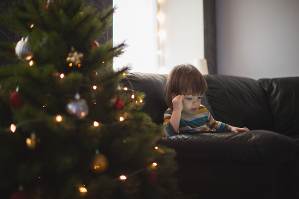 Petit garçon aux cheveux longs regarde une vidéo sur le téléphone assis sur le canapé près du sapin de Noël. Un enfant joue au téléphone pour la nouvelle année
 - Photo, image