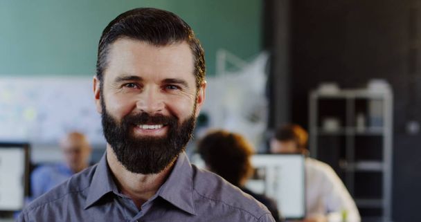 Крупный план привлекательного молодого человека с бородой, смотрящего и улыбающегося в камеру. Размытый офис с людьми на компьютерном фоне. Внутрь. Портрет
 - Фото, изображение