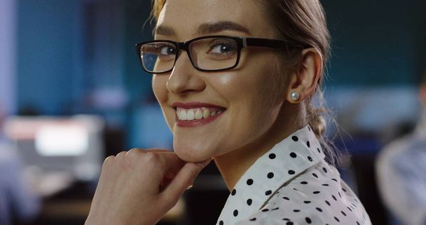 Close up of beautiful woman in glasses looking at office space behind her, than turning the head to the camera and smiling sincerely. Late in the evening. Indoor. Portrait - Zdjęcie, obraz