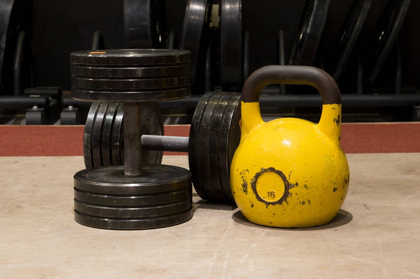 Two old and used gym black metal dumbbells with big yellow kettlebell on a wooden floor - Foto, Bild