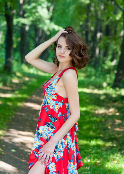 Young brunette girl in red dress posing on alley in summer park against trees - Fotó, kép