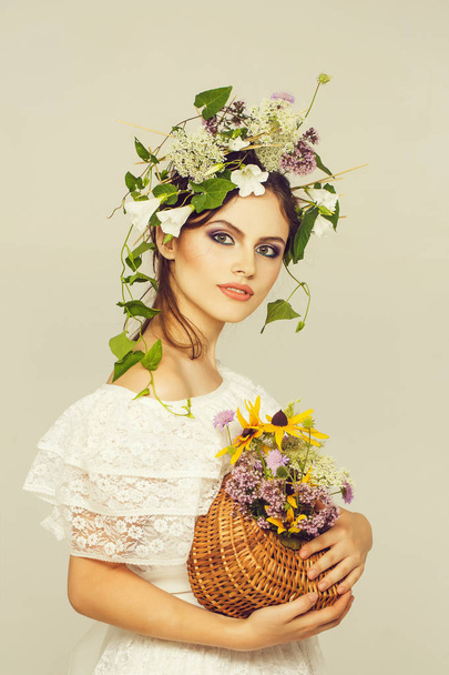 sexy girl or woman with fashionable makeup on pretty face and flower wreath on long brunette hair in wedding dress holding basket with bouquet in studio isolated on white background - Photo, image