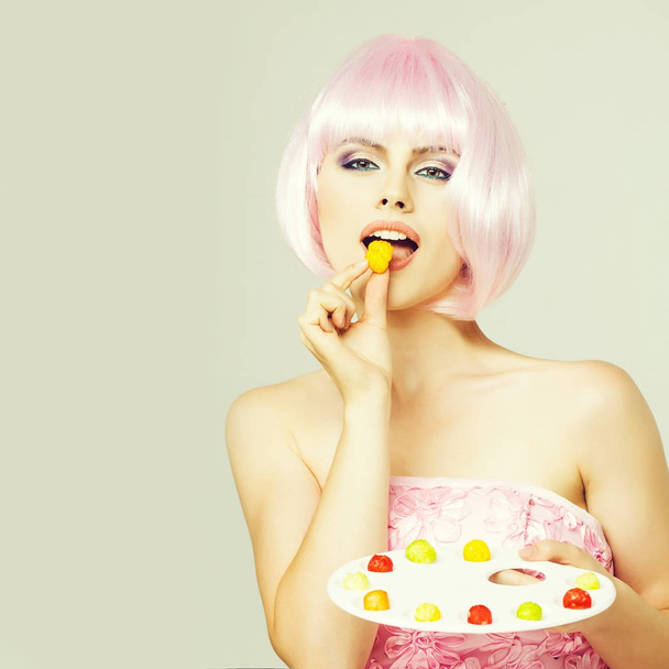 sexy glamour girl or woman with fashionable makeup on pretty face and short hairstyle or pink wig in dress eating colorful marmalade sweets on paint palette in studio on grey background, copy space - Foto, Bild