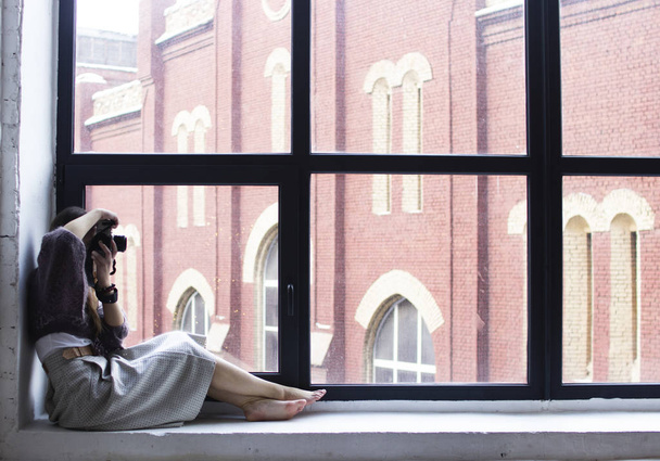 a young woman sits on a windowsill with large windows and takes  - 写真・画像