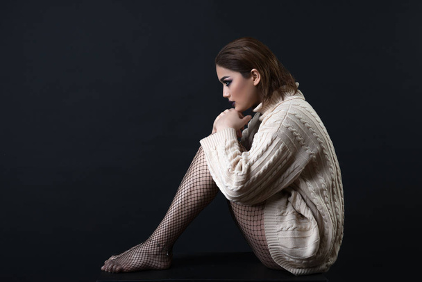 Nuori kaunis nainen yllään villapaita paljain jaloin istuu musta tausta
 - Valokuva, kuva