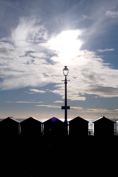 une ligne de 5 cabanes de plage et un lampadaire silhoutted noir par le soleil brille directement à la caméra à travers un ciel bleu et blanc dramatique, la mer scintille derrière les cabanes de plage
,   - Photo, image