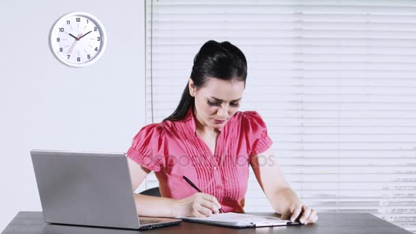 Young Caucasian businesswoman working on desk while writing on a clipboard with a laptop computer in the office - Video