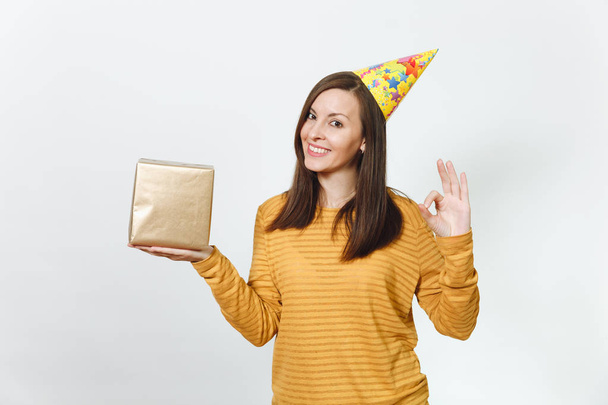 Cheerful caucasian young happy woman in yellow clothes, birthday party hat holding golden gift box with present, celebrating holiday, showing OK gesture on white background isolated for advertisement. - Photo, Image