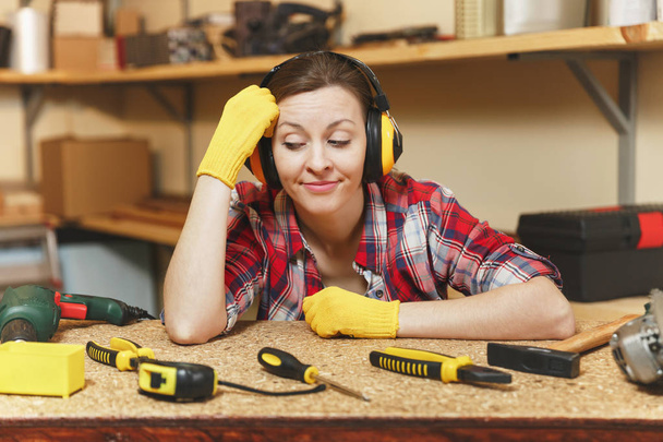 Ratlos ratlose junge Frau in kariertem Hemd, grauem T-Shirt, lärmgedämmten Kopfhörern, gelben Handschuhen, die in der Tischlerei am Holztisch mit Holzstück arbeiten, verschiedene Werkzeuge. - Foto, Bild