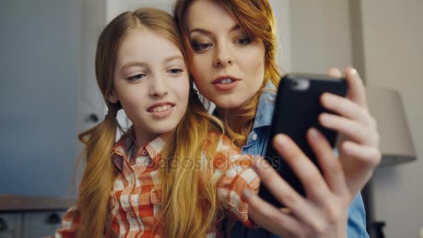 Close up of the beautiful daughter and mother sitting close to each other and making selfies on the black smartphone in the room at home. Indoors. Portrait - Footage, Video