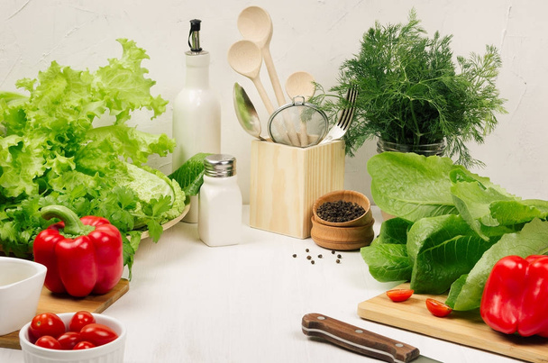 Kitchen white interior with raw fresh green salad, red cherry tomatoes, kitchenware on soft white wood table, copy space. Spring vitamin dieting food. - Valokuva, kuva
