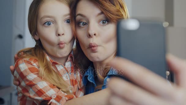 Close up of the smiling mother hugging young daughter and making funny selfies on the smartphone while grimacing. Portrait. Inside - Materiał filmowy, wideo