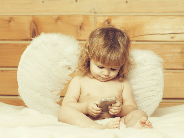 Cute happy beautiful playful child boy with wet hair sitting in hothouse bath white fluffy towel naked indoor on wooden background in feathered angel wings playing on mobile phone, horizontal picture - Foto, Bild