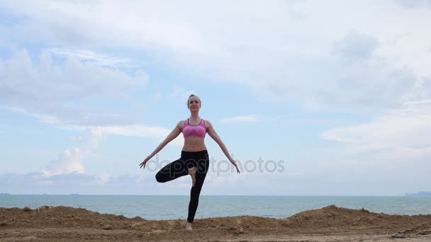 Girl practices yoga fitness exercise near the ocean beach in tree pose - 映像、動画