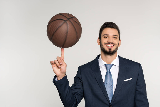 young businessman holding basketball ball on finger and smiling at camera isolated on grey   - Foto, immagini