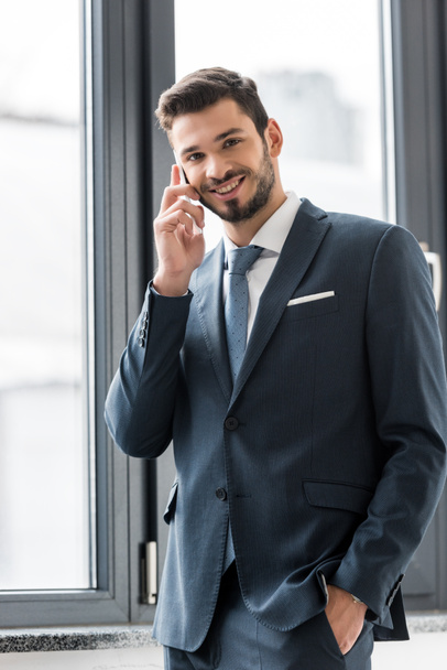 young businessman talking on smartphone and smiling at camera in office - Фото, зображення