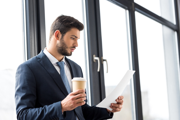 young businessman holding coffee to go and reading papers in office - Photo, image