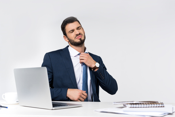 joven empresario sobrecargado de trabajo con los ojos cerrados ajustando la corbata mientras está sentado en el lugar de trabajo
  - Foto, imagen