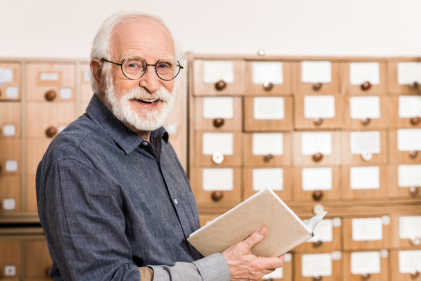 uśmiechający się starszy mężczyzna archiwista prowadzące księgę i patrząc na kamery - Zdjęcie, obraz
